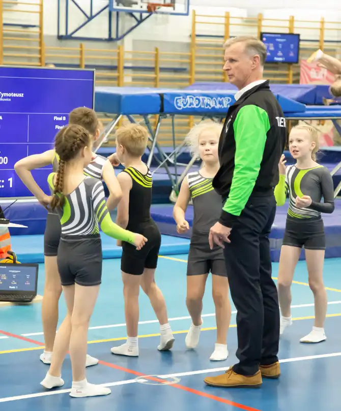 Trener w klubowym stroju czuwa nad grupą dzieci w klubowych strojach, w tle hala zawodów