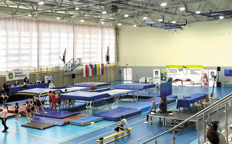 Wnętrze dużej, jasnej sali gimnastycznej wypełnione trampolinami i materacami, pośród nich zawodnicy, w tle na drabinkach wiszą flagi Polski, Słowacji, Czech i Ukrainy.