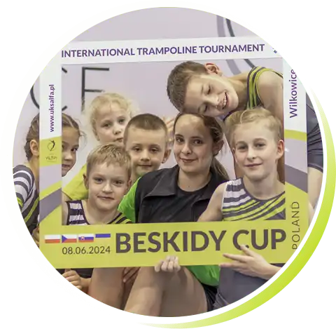 Grupa dzieci w różnym wieku, chłopcy i dziewczynki w klubowych strojach uśmiechnięci pozują do zdjęcia z foto-ramką międzynarodowych zawodów "Beskidy Cup Wilkowice"