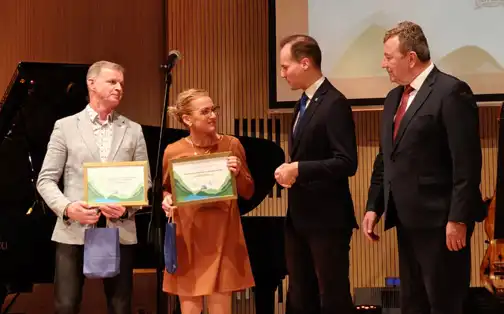 Janusz Kruk na scenie uroczystej gali z nagrodą, obok niego inna nagrodzona osoba i dwie osoby wręczające nagrody.