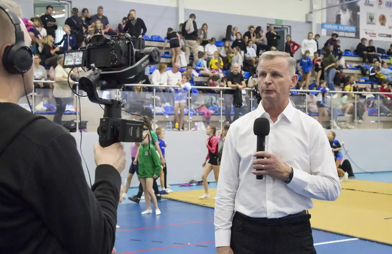 Organizator zawodów z mikrofonem, przed nim operator kamery ze sprzętem rejestrującym obraz i dźwięk, w tle hala sportowa z trybunami wypełnionymi kibicami.