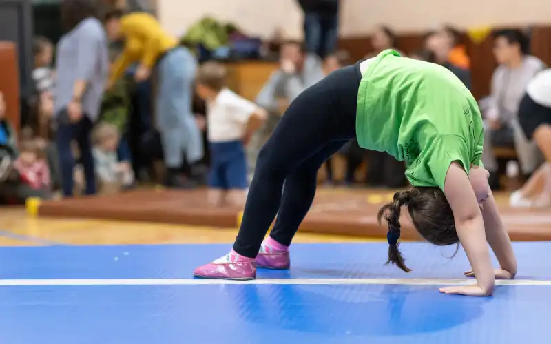 Na pierwszym planie mała dziewczynka robiąca "mostek" na ścieżce gimnastycznej, w tle grupa kibiców na sali gimnastycznej.