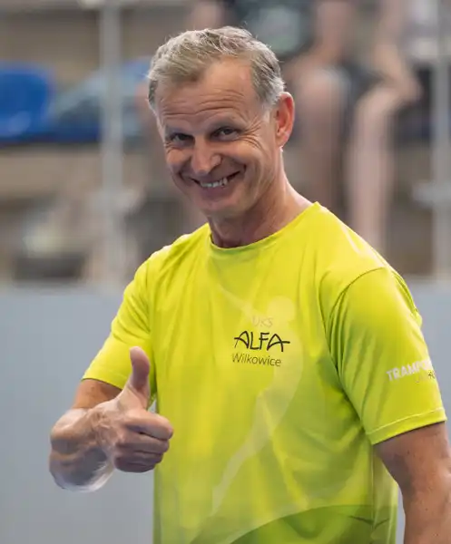 Roześmiany trener w klubowej koszulce UKS ALFA Wilkowice pokazuje "ok", w tle trybuny hali sportowej.