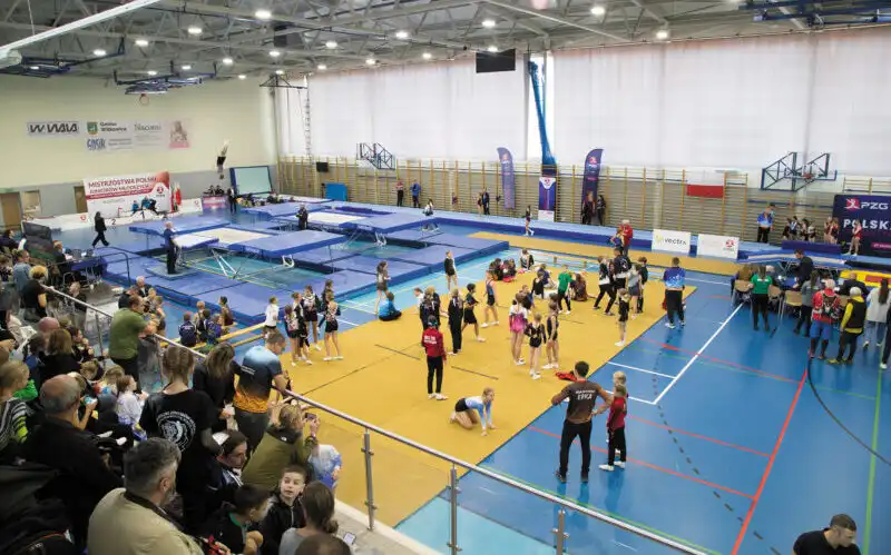 Wnętrze hali sportowej wypełnione dwoma trampolinami z przystawkami, materacami i planszą treningową. Na sali znajduje się dużo zawodników. W narożniku zdjęcia na pierwszym planie widać wypełnione kibicami trybuny.