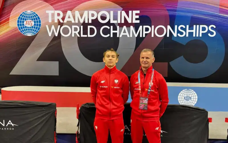 Trener z zawodnikiem, w dresach reprezentacji Polski, stoją na tle ścianki z napisem "World Trampoline Championschips 2025"