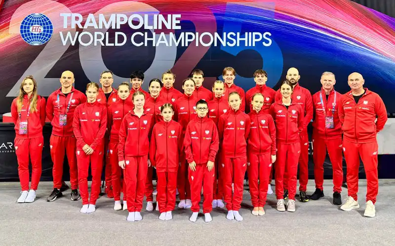 Zdjęcie grupowe - zawodnicy w reprezentacyjnych strojach Polskiej Kadry Narodowej stoją przed ogromnym banerem Mistrzostw Świata "Trampoline World Championschips 2025" Wśród nich zawodnik Filip Procner i trener Janusz Kruk.