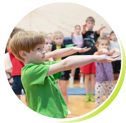 Grupa dzieci na treningu ćwiczy równowagę. Skupiony chłopczyk na pierwszym planie wyciąga przed siebie równo ułożone ręce.