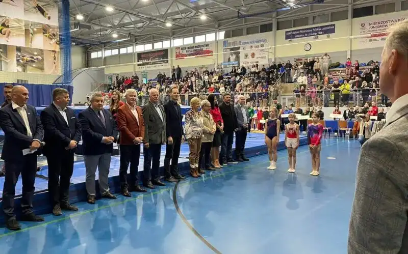 Grono oficjeli oraz poczet flagowy zawodników podczas otwarcia zawodów, w tle hala zawodów z trampolinami.