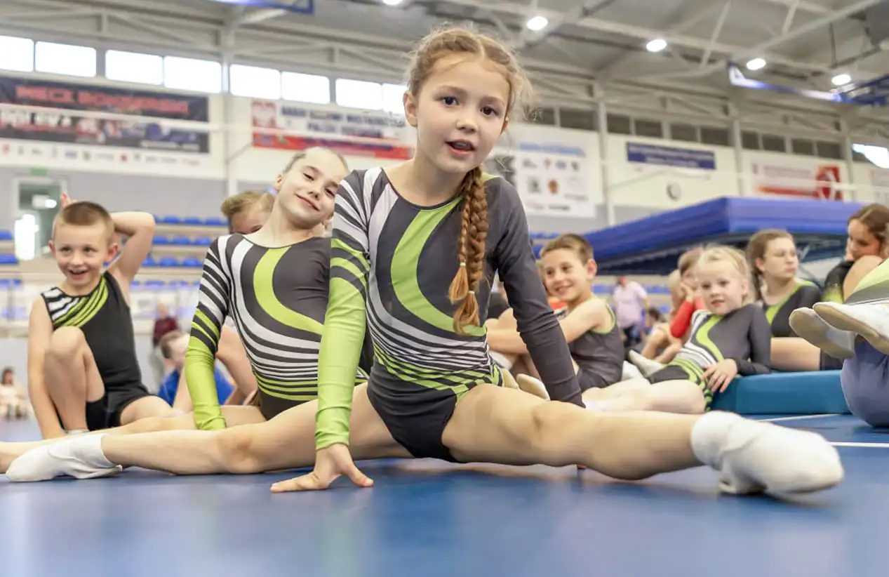 Grupa uśmiechniętych dzieci w strojach klubowych, na pierwszym planie dziewczynka robiąca szpagat, w tle hala sportowa z trybunami i trampolinami.