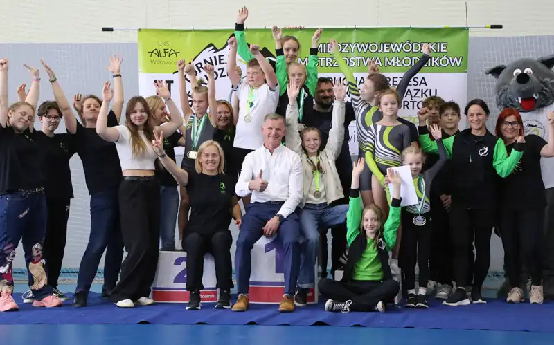 Grupa osób w różnym wieku, część w strojach klubowych, radośnie machają stojąc na podium na tle baneru zawodów.